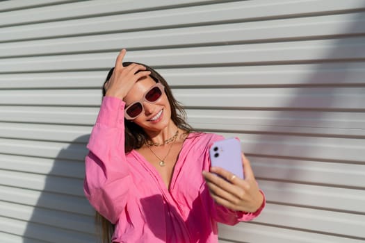 Young beautiful brunette in a pink shirt, neck jewelry, necklace, trendy sunglasses on the background of a light garage fence, takes a selfie on the phone, laughs, smiles