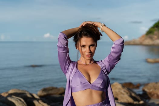 Young beautiful woman in a romantic mood, lilac silk clothes, on the beach against the backdrop of the sea and stones at sunset