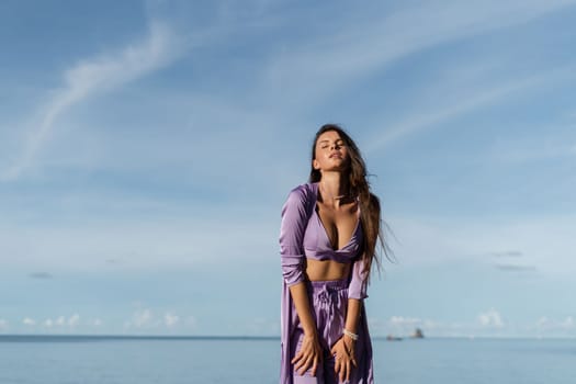 Young beautiful woman in a romantic mood, lilac silk clothes, on the beach against the backdrop of the sea and stones at sunset