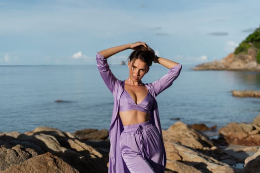 Young beautiful woman in a romantic mood, lilac silk clothes, on the beach against the backdrop of the sea and stones at sunset