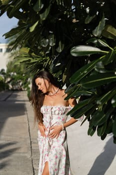 Young beautiful woman in a romantic dress with a floral print, and a pearl necklace bracelet, against the backdrop of tropical leaves