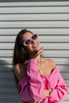 Young beautiful brunette in a pink shirt, neck jewelry, necklace, trendy sunglasses on the background of a light garage door fence