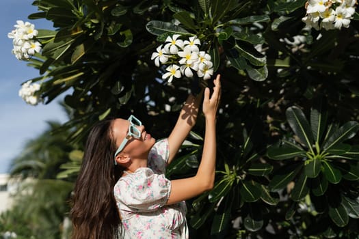 Young beautiful woman in a romantic dress with a floral print, sunglasses and a pearl necklace bracelet, against the backdrop of tropical leaves