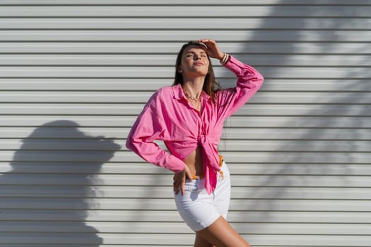 Young beautiful brunette woman in a pink shirt and white denim shorts against the background of a light garage door fence