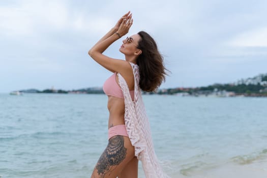 Stylish young woman with voluminous hair in a bikini and lace pareo posing on a tropical beach, slim, tanned, athletic, sunglasses