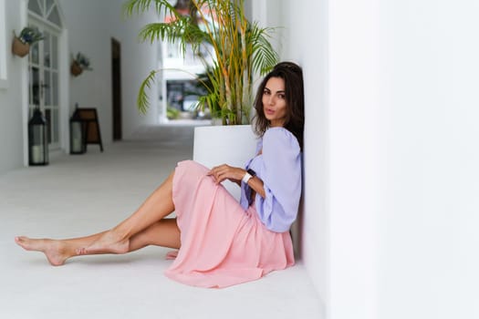 Stylish young woman with voluminous hair in a trendy long sleeve crop top and a pink skirt posing against a white wall and a tropical bush, wearing a smart watch