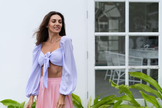 Pretty young stylish woman in trendy purple crop top long sleeve , pink skirt, isolated, white wall, tropical leaves natural day light..