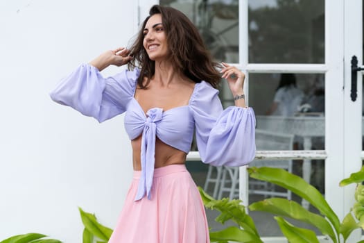 Pretty young stylish woman in trendy purple crop top long sleeve , pink skirt, isolated, white wall, tropical leaves natural day light..