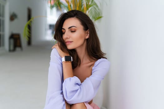 Stylish young woman with voluminous hair in a trendy long sleeve crop top and a pink skirt posing against a white wall and a tropical bush, wearing a smart watch