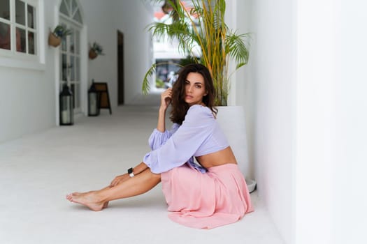 Stylish young woman with voluminous hair in a trendy long sleeve crop top and a pink skirt posing against a white wall and a tropical bush, wearing a smart watch