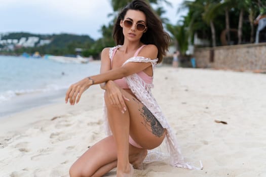 Stylish young woman with voluminous hair in a bikini and lace pareo posing on a tropical beach, slim, tanned, athletic, sunglasses