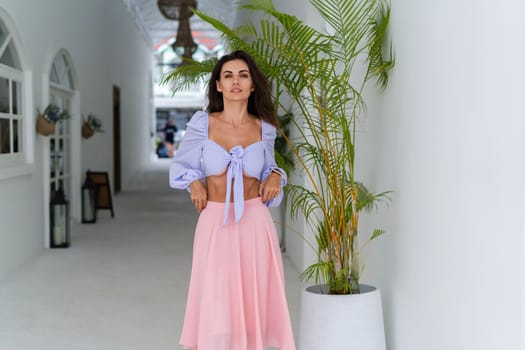 Stylish young woman with voluminous hair in a trendy long sleeve crop top and a pink skirt posing against a white wall and a tropical bush, wearing a smart watch