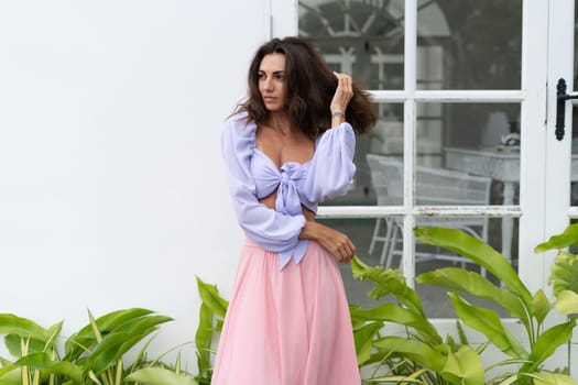 Pretty young stylish woman in trendy purple crop top long sleeve , pink skirt, isolated, white wall, tropical leaves natural day light..