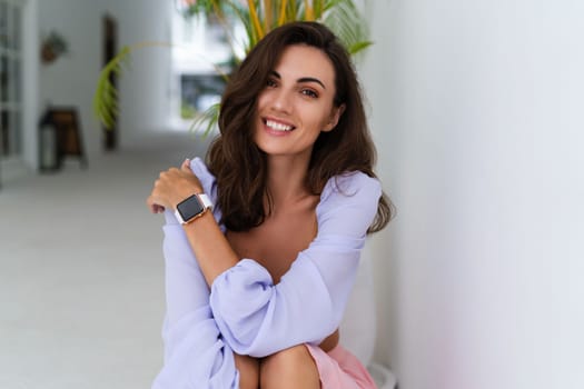 Stylish young woman with voluminous hair in a trendy long sleeve crop top and a pink skirt posing against a white wall and a tropical bush, wearing a smart watch