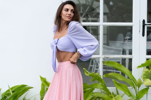 Pretty young stylish woman in trendy purple crop top long sleeve , pink skirt, isolated, white wall, tropical leaves natural day light..