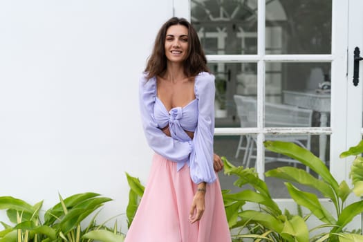 Pretty young stylish woman in trendy purple crop top long sleeve , pink skirt, isolated, white wall, tropical leaves natural day light..