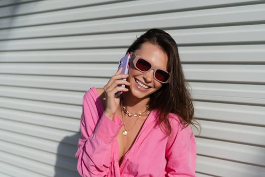 Young beautiful brunette woman in a pink shirt, neck jewelry, necklace, trendy sunglasses on the background of a light garage fence, calls the phone, laughs, smiles