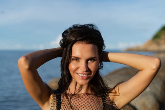 A young beautiful tanned woman against the backdrop of the sea at sunset in a black swimsuit and a mesh dress, laughs and smiles with a snow-white smile