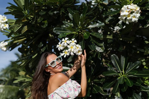 Young beautiful woman in a romantic dress with a floral print, sunglasses and a pearl necklace bracelet, against the backdrop of tropical leaves