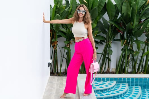 Stylish fit fashion women in bright pink wide leg pants holding bag trendy mint sunglasses posing at luxury tropical villa by pool outdoor natural day light