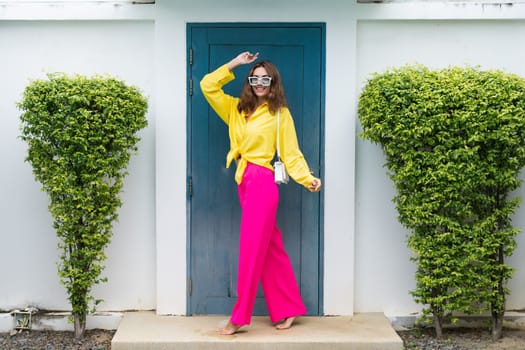 Stylish fit fashion women in bright pink wide leg pants and yellow shirt holding bag trendy mint sunglasses posing at luxury tropical villa by pool outdoor natural day light