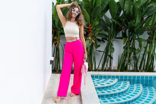 Stylish fit fashion women in bright pink wide leg pants holding bag trendy mint sunglasses posing at luxury tropical villa by pool outdoor natural day light