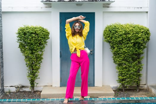 Stylish fit fashion women in bright pink wide leg pants and yellow shirt holding bag trendy mint sunglasses posing at luxury tropical villa by pool outdoor natural day light