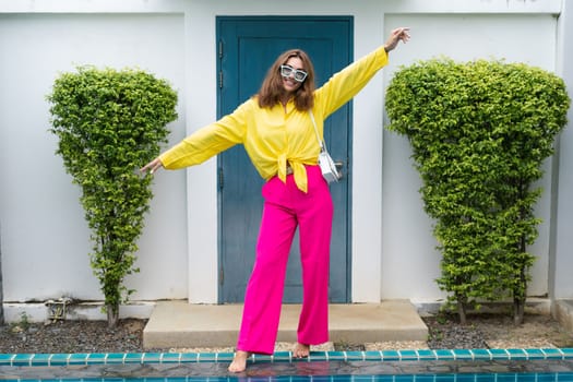 Stylish fit fashion women in bright pink wide leg pants and yellow shirt holding bag trendy mint sunglasses posing at luxury tropical villa by pool outdoor natural day light