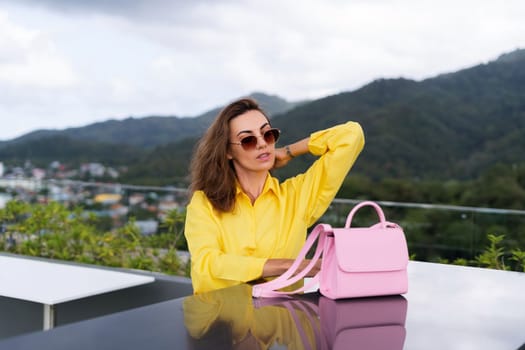 Stylish fit fashion women in bright yellow shirt trendy sunglasses posing at rooftop terrace tropical view outdoor with pink bag