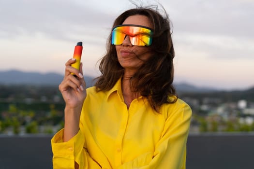 Stylish fit fashion women in bright yellow shirt trendy shield visor rainbow sunglasses posing at rooftop terrace tropical view sunset smoking vape pod enjoying puffs