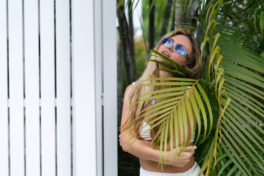 Beautiful young blonde woman in a top with long hair, natural beauty, daylight, in blue glasses, posing against the backdrop of a wooden fence and palm leaves