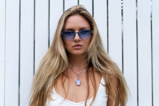 Beautiful young blonde woman in a top with long hair and smooth clean soft skin, natural beauty, daylight, wearing blue glasses, posing against the backdrop of a wooden fence