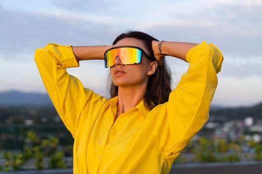 Stylish fit fashion women in bright yellow shirt trendy shield visor rainbow sunglasses posing at rooftop terrace tropical view outdoor sunset warm light