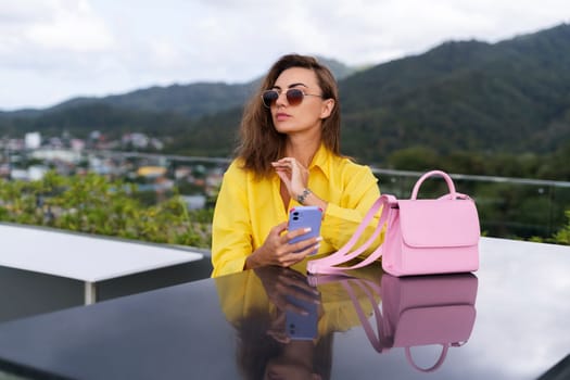 Stylish fit fashion women in bright yellow shirt trendy sunglasses posing at rooftop terrace tropical view outdoor holding mobile phone