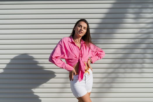 Young beautiful brunette woman in a pink shirt and white denim shorts against the background of a light garage door fence