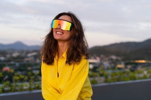 Stylish fit fashion women in bright yellow shirt trendy shield visor rainbow sunglasses posing at rooftop terrace tropical view outdoor sunset warm light