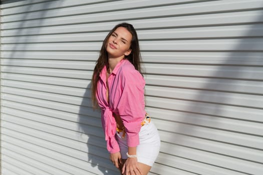 Young beautiful brunette woman in a pink shirt and white denim shorts against the background of a light garage door fence