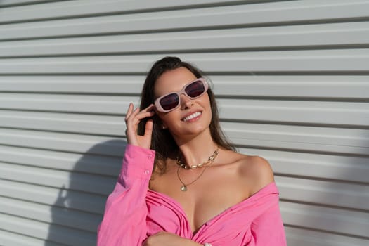 Young beautiful brunette in a pink shirt, neck jewelry, necklace, trendy sunglasses on the background of a light garage door fence