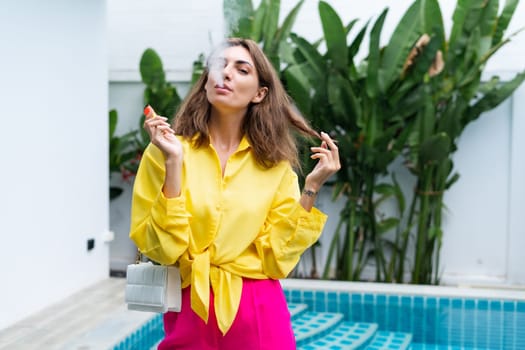 Stylish fit fashion women in bright pink pants holding bag trendy posing at luxury tropical villa by pool outdoor natural day light smoking vape pod enjoying puffs