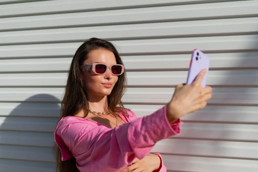 Young beautiful brunette in a pink shirt, neck jewelry, necklace, trendy sunglasses on the background of a light garage fence, takes a selfie on the phone, laughs, smiles