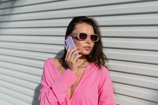 Young beautiful brunette woman in a pink shirt, neck jewelry, necklace, trendy sunglasses on the background of a light garage fence, calls the phone, laughs, smiles