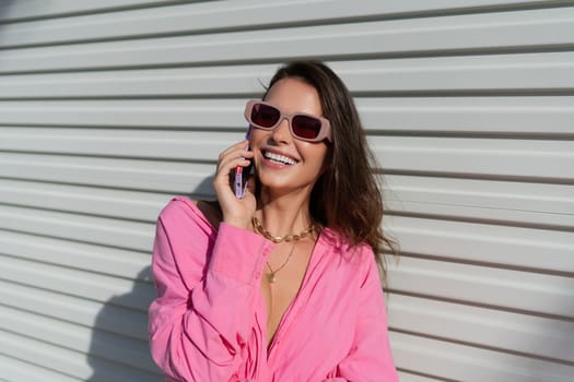 Young beautiful brunette woman in a pink shirt, neck jewelry, necklace, trendy sunglasses on the background of a light garage fence, calls the phone, laughs, smiles