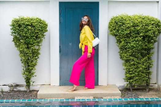 Stylish fit fashion women in bright pink wide leg pants and yellow shirt holding bag trendy mint sunglasses posing at luxury tropical villa by pool outdoor natural day light