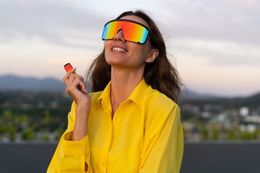 Stylish fit fashion women in bright yellow shirt trendy shield visor rainbow sunglasses posing at rooftop terrace tropical view sunset smoking vape pod enjoying puffs