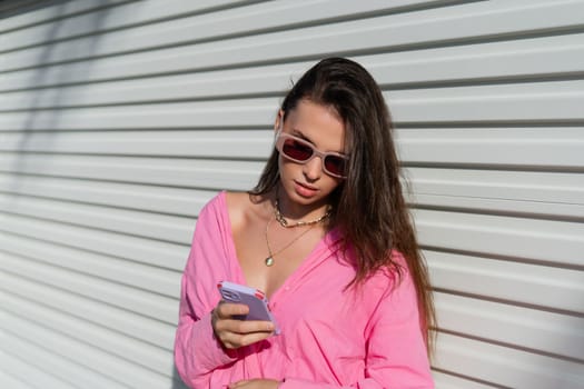 Young beautiful brunette in a pink shirt, neck jewelry, necklace, trendy sunglasses on the background of a light garage fence, looks at the phone screen