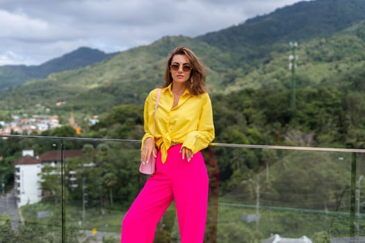 Stylish fit fashion women in bright pink wide leg pants and yellow shirt holding bag trendy sunglasses posing at rooftop terrace tropical view outdoor natural day light