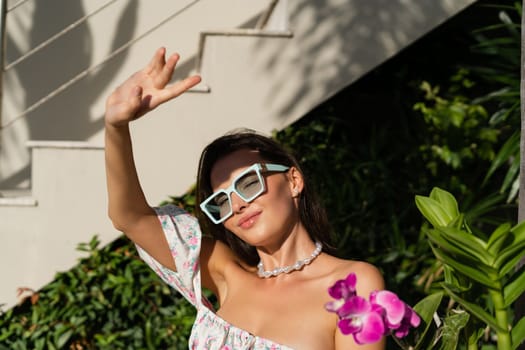 Young beautiful woman in a romantic dress with a floral print, sunglasses and a pearl necklace bracelet, against the backdrop of tropical leaves