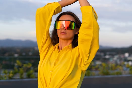 Stylish fit fashion women in bright yellow shirt trendy shield visor rainbow sunglasses posing at rooftop terrace tropical view outdoor sunset warm light