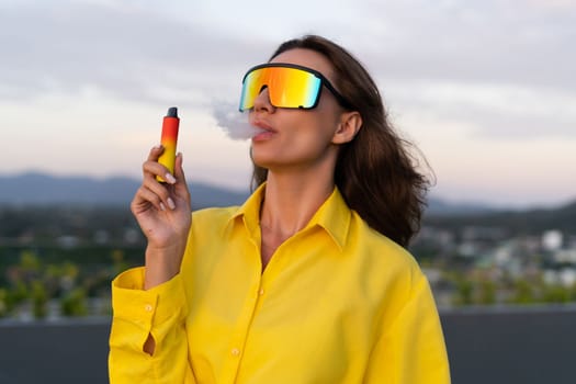 Stylish fit fashion women in bright yellow shirt trendy shield visor rainbow sunglasses posing at rooftop terrace tropical view sunset smoking vape pod enjoying puffs