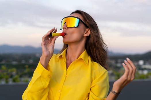 Stylish fit fashion women in bright yellow shirt trendy shield visor rainbow sunglasses posing at rooftop terrace tropical view sunset smoking vape pod enjoying puffs